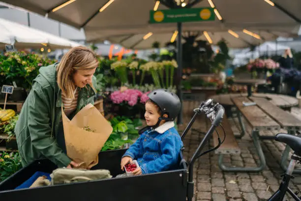 Photo of a boy riding in a cargo bike, having a healthy snack while grocery shopping with his mother at a local market.