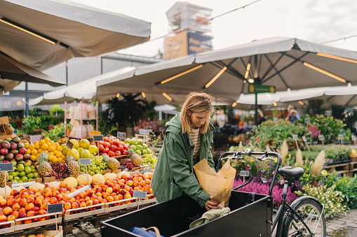 Photo of a young woman loading groceries into her cargo bike while grocery shopping at a local food market.