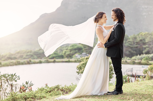 Wedding, wind and a couple, veil waving in beautiful natural landscape. Nature, love and a happy bride and groom standing in park together. Luxury venue, man and woman at outdoor marriage ceremony.