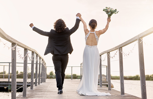 Couple, walking and wedding, celebration of love with hands in sky and bridge at lake or dam together. Man, bride with bouquet of flowers and walk by river, happy on day of marriage in summer