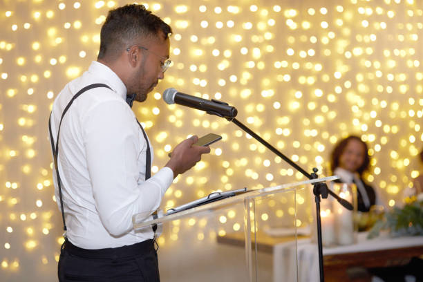 istock Speech, wedding and microphone with a man reading from his phone during a marriage ceremony or celebration event. Talking, toast and tradition with a handsome young male speaking at an indoor venue 1432006158