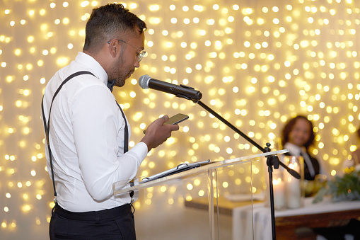 Speech, wedding and microphone with a man reading from his phone during a marriage ceremony or celebration event. Talking, toast and tradition with a handsome young male speaking at an indoor venue