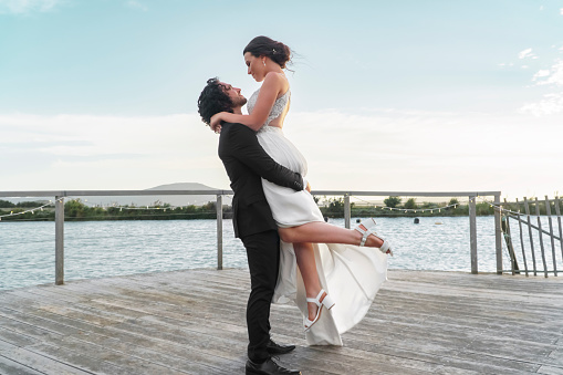 Wedding by lake, man and woman together on deck in nature and love. Beautiful couple relax by the water in the natural afternoon sun, white dress of bride and black wedding suit contrast in sunshine