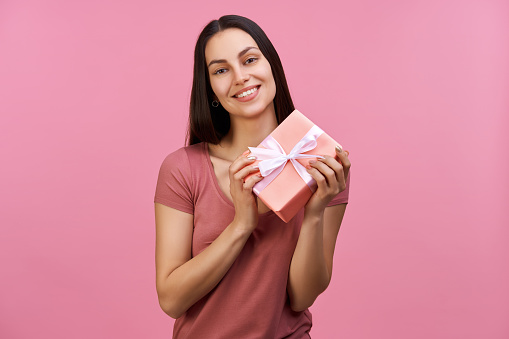 Studio shot of happy smiling attractive young brunette woman holding gift box, isolated over pastel pink background.