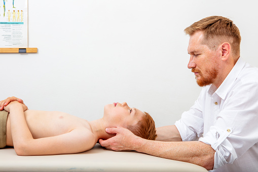 Male osteopath (chiropractor) treating neck problem of a boy lying down on a massage bed. Can illustrate the concept of Osteopathy, Alternative medicine, Physiotherapy, pain relief. Space for copy.