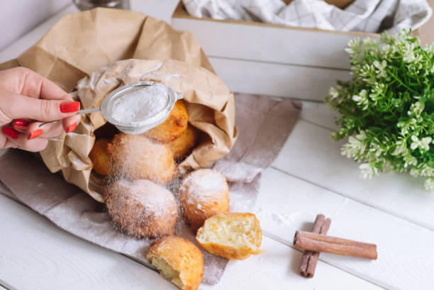 Female hand sprinkles powdered sugar on donuts. Cottage cheese round donuts pile covered up with sugar powder, big pile in a paper bag. Whole dough balls no holes. stock photo