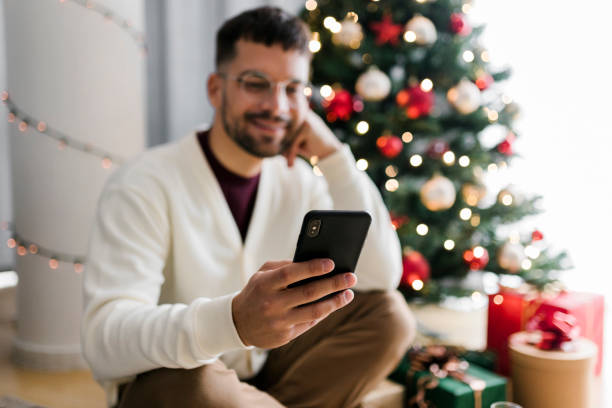 istock Young man sitting by Christmas tree and using his smart phone 1432013438