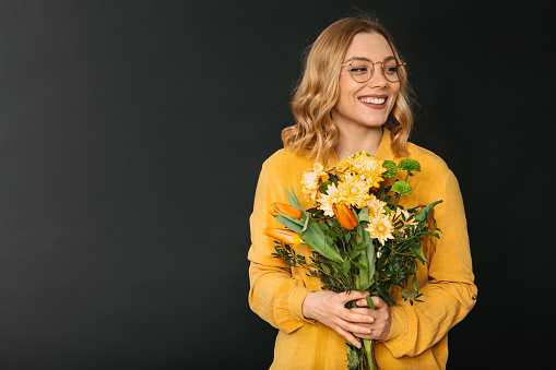 Studio shot of a young blonde woman in a yellow shirt wearing eyeglasses holding a bouquet of colorful flowers