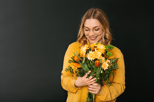Studio shot of a young blonde woman in a yellow shirt wearing eyeglasses holding a bouquet of colorful flowers