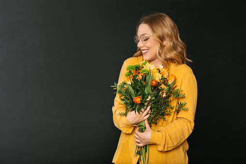 Studio shot of a young blonde woman in a yellow shirt wearing eyeglasses holding a bouquet of colorful flowers