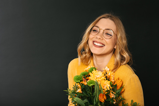 Studio shot of a young blonde woman in yellow shirt wearing eyeglasses holding a bouquet of colourful flowers