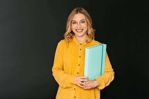 A young and beautiful blonde woman holding a folder on dark background in yellow shirt