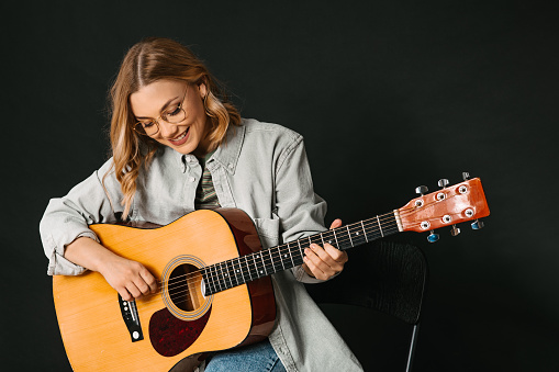 A young blonde woman in shirt playing a guitar and sitting on chair