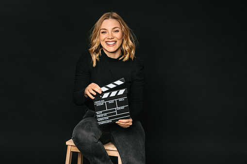 A young blonde female director holding a film slate while sitting on a high black chair and smiling