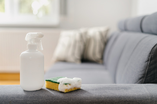 detergent spray in white bottle with blank space and scrubber sponge with foam on grey sofa in living room with blurred background