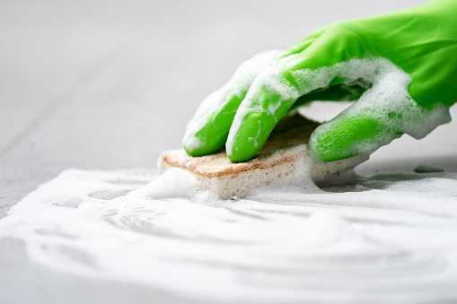 female hand in green rubber glove wash floor surface with soap foam and sponge in apartment room, cropped shot, cleaning service concept