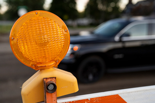An orange colored construction safety light sits on top of a white and orange safety barricade, a black car passes in the background.  Room for text at the side of the image.