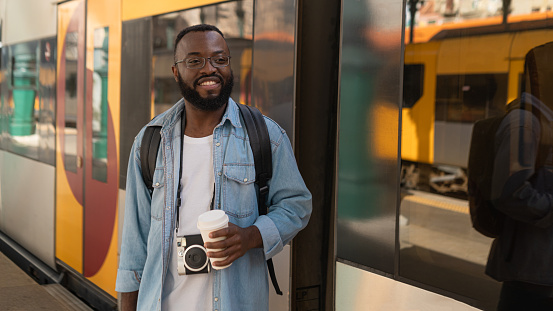 Smiling african american man with coffee at european terminal train station. Tourist travel by train on vacation time holiday weekend trip.