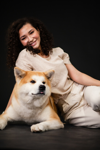 happy young  Woman hugging Akita inu Dog. Japanese akita dog and Curly brunette  posing together on dark background, pets and leisure concept
