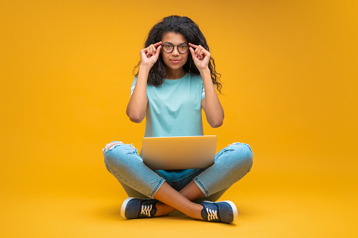 Studio portrait of cute young african american student girl in trendy spectacles, sitting with laptop and looking directly to the camera, isolated over bright orange yellow background.