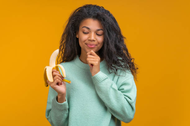 ritratto in studio di una ragazza bruna sorridente vestita casualmente che va a mangiare una banana fresca matura che tiene in mano, isolata su uno sfondo giallo colorato brillante. - pilled foto e immagini stock