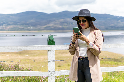 A Latin young adult woman with glasses, coat and hat is looking her phone near a lake