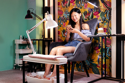 smiling young woman using a mobile phone while sitting in a beauty salon chair during her pedicure treatment, wellness and body care concept
