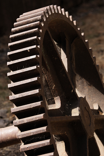 Old rusted gear, very close up, very tightly cropped, set slanting toward the left in this vertical composition.  The deeply set teeth arch downward in the left half of the frame from top center to bottom.  A small portion of the gear shaft appears at bottom left, extending out of frame.  Strong sunlight and shadows help add drama to this image in mostly rust-brown colors.