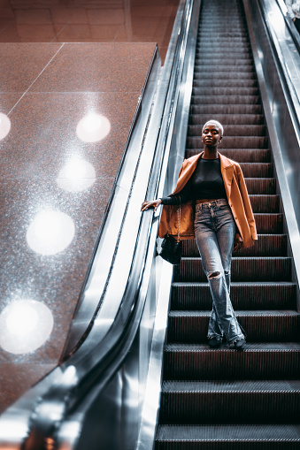 A vertical shot of a young elegant black female descending to an underground passage using a moving staircase; a cute black woman in a coat is going down on the escalator to the subway station depot