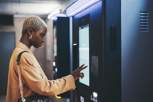 A side view of a young beautiful elegant black woman with painted white very short hair paying for service underground parking or buying a subway or train ticket using an electronic self-service kiosk
