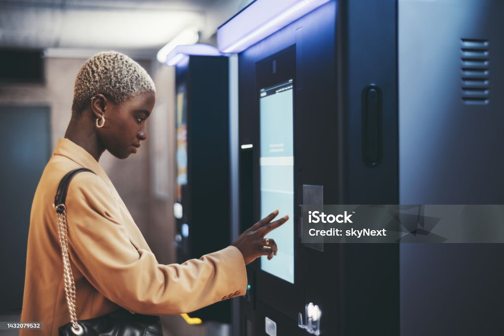 African girl using ticket terminal A side view of a young beautiful elegant black woman with painted white very short hair paying for service underground parking or buying a subway or train ticket using an electronic self-service kiosk ATM Stock Photo