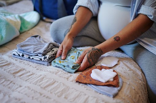 Pregnant, woman and baby, clothes on bed with mother fold laundry in a bedroom for newborn. Love, future and planning by excited lady choosing outfit for birth of child, preparation and hands closeup