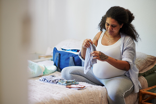 Pregnant mom, packing clothes for baby birth in bag and ready for pregnancy emergency hospital trip. Woman folding child clothing, on bed in bedroom and for doctor visit for health checkup at clinic