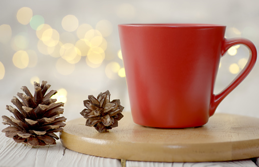 Red mug on wooden board with pine cones against white bokeh orb background