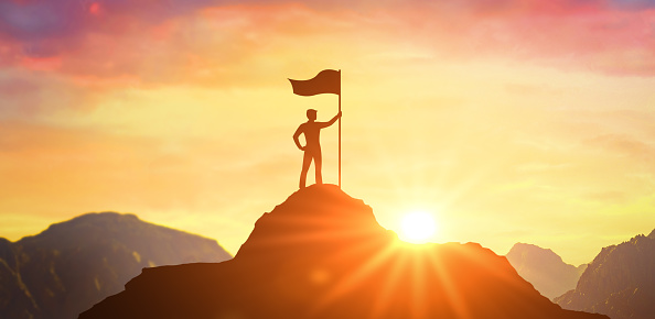 Silhouette of businessman holding flag on top mountain, sky and sun light background. Business success and goal concept. Man silhouette on mountain top. Concept of success