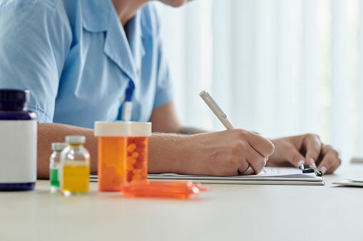 Close-up of young nurse prescribing medicine for patient while sitting at table with bottles of pills