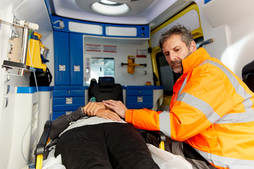 Photo with copy space of serious male paramedic seated at the side of an injured woman who is outstretched on a gurney; EMS vehicle equipment seen in background. Copy space