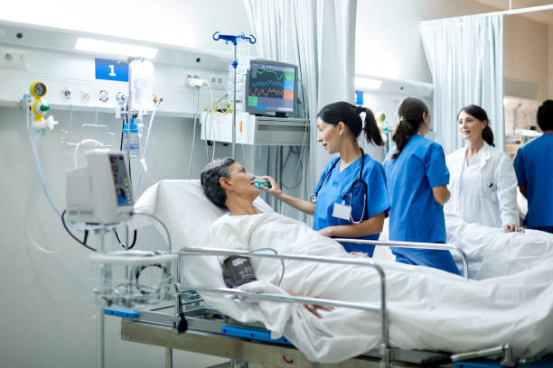 Nurse adjusting medical equipment while speaking with female patient in open plan hospital ward with patients and medical personnel stock photo