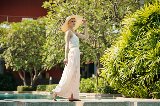 Elegant young woman in dress and hat walking along the outdoor pool at resort