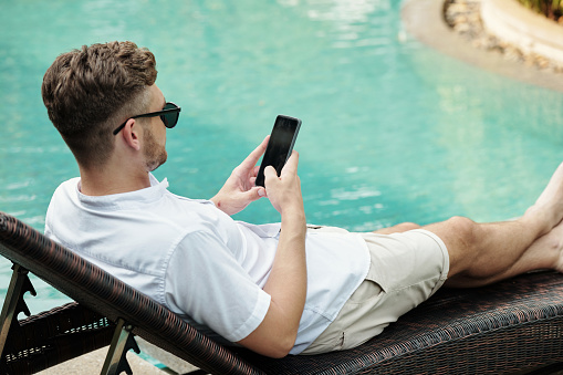 Young man in sunglasses using mobile phone while resting on sunbed near the outdoor pool