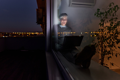Happy senior entrepreneur working on a computer by the window at night. The view is through glass.