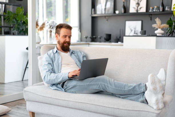 istock Young bearded man sitting on sofa at home using laptop computer 1432124590