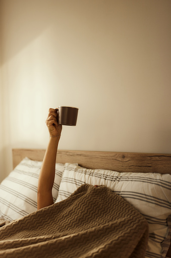 Unrecognizable person covered with blanket raising her hand in a bed while wanting a cup of morning coffee. Copy space.