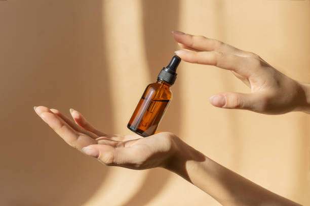 bottle of serum in women's hands. glass bottle with dropper cap in women's hands. amber glass container with dropper lid for cosmetic products on brown background in sunlight - druppelfles stockfoto's en -beelden