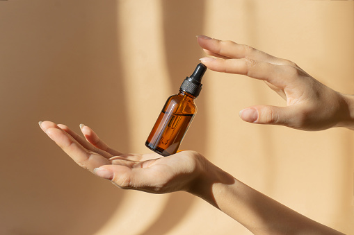 Bottle of serum in women's hands. Glass bottle with dropper cap in women's hands. Amber glass container with dropper lid for cosmetic products on brown background in sunlight.