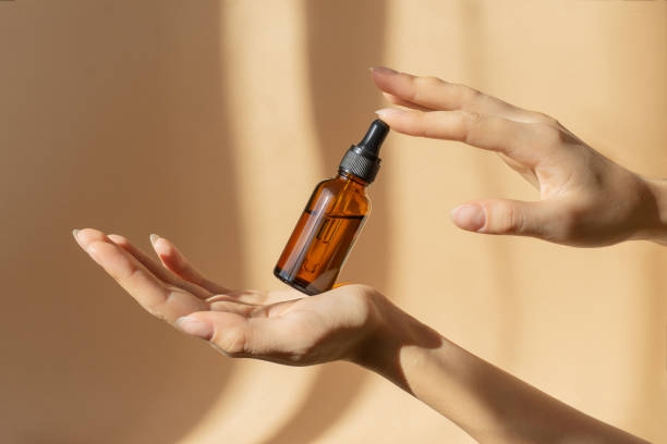 Bottle of serum in women's hands. Glass bottle with dropper cap in women's hands. Amber glass container with dropper lid for cosmetic products on brown background in sunlight Bottle of serum in women's hands. Glass bottle with dropper cap in women's hands. Amber glass container with dropper lid for cosmetic products on brown background in sunlight. pipette photos stock pictures, royalty-free photos & images