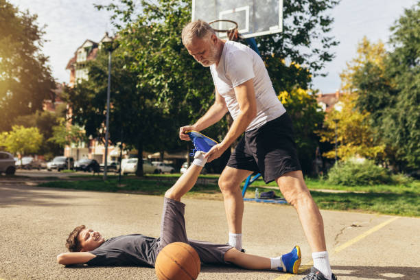 padre che aiuta il ragazzo con un trauma alle gambe dopo aver giocato a basket. infortunio sportivo. - athlete coach basketball male foto e immagini stock