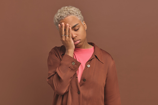 Young exhausted ethnic African American woman closes eyes leans on hand suffers from chronic fatigue or problems associated with lack of sleep and overworking stands on brown studio background