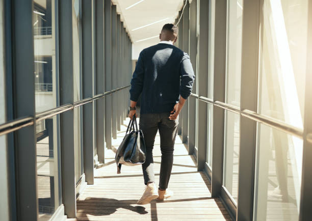 Businessman at airport walking for work travel and male african morning commute to city. Transport service from airplane terminal to hotel, rear view of modern worker carrying hand luggage bag trip. hombre de negocios en el aeropuerto caminando para viajes de trabajo y hombre africano por la mañana viaje a la ciudad. servicio de transporte desde la terminal del avión hasta el hotel, vista trasera del trabajador moderno que lleva equipaje de mano de