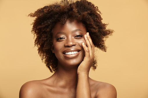 Beautiful headshot portrait of hilarious African American ethnicity woman with charming broad smile having a healthy clean skin standing on a beige isolated background. Adult dark skinned girl with a curly afro style hair.